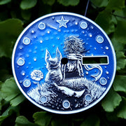 This image shows a dial with the little prince sitting on an asteroid. The little prince is dressed in a suit and scarf. A fox sits on an asteroid next to a rose. The image has a round shape and a blue background.
