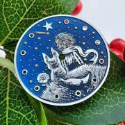 This image shows a dial with the little prince sitting on an asteroid. The little prince is dressed in a suit and scarf. A fox sits on an asteroid next to a rose. The image has a round shape and a blue background.
